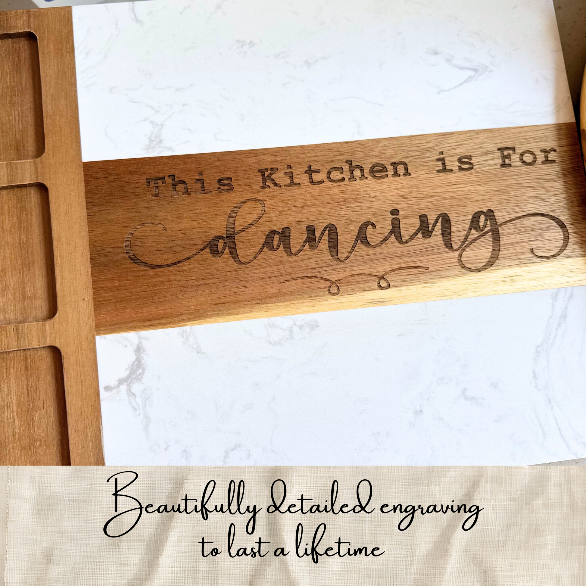 A close-up of a wooden kitchen utensil or board engraved with the phrase 'This Kitchen is For dancing' in dark lettering, placed over a white marble-patterned surface. A beige fabric banner at the bottom reads 'Beautifully detailed engraving to last a lifetime.