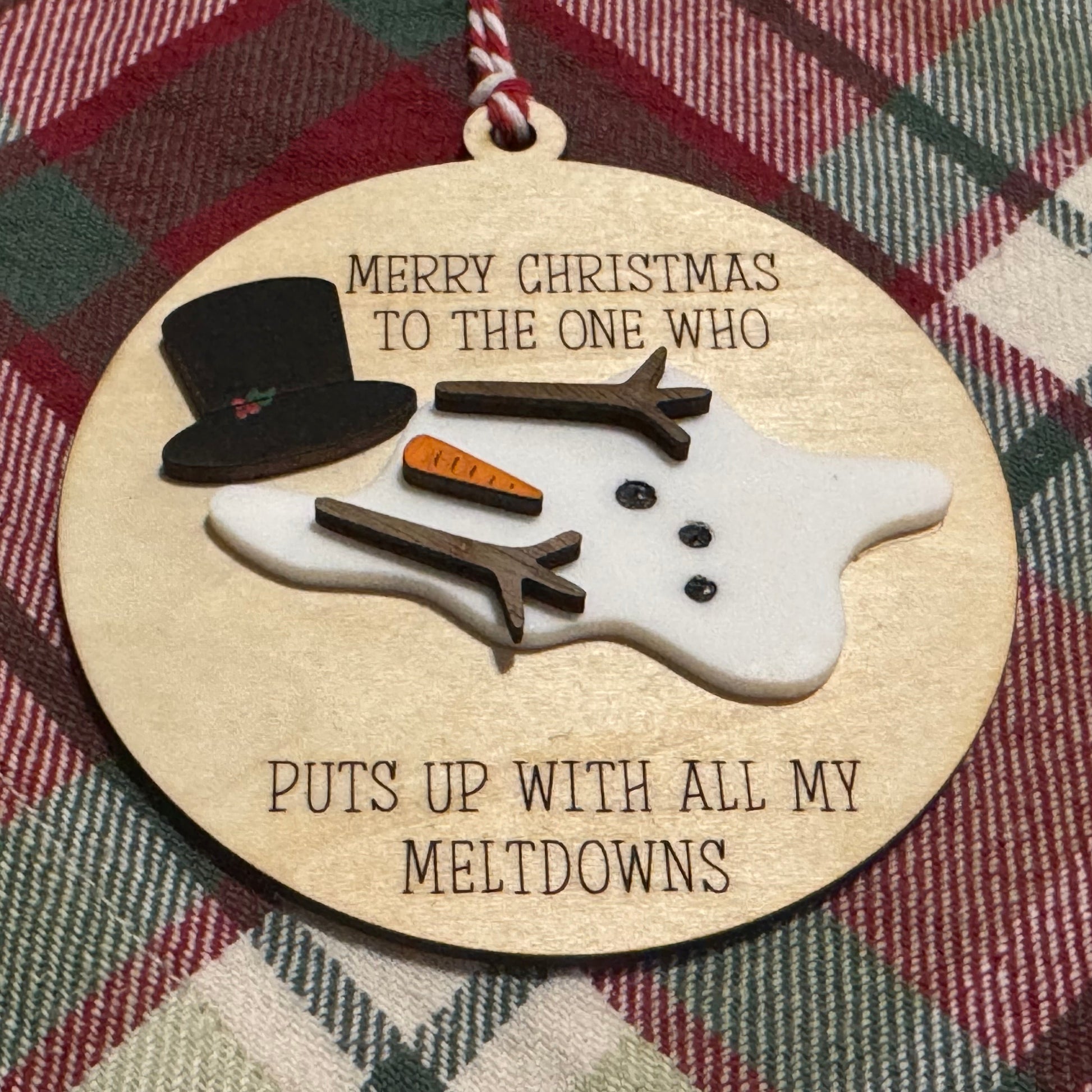 Decorative Christmas ornament with a snowman design and humorous text on a plaid fabric background.