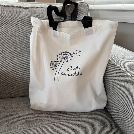 tote bag with black dandelion design and 'Just breathe' text on a gray couch.