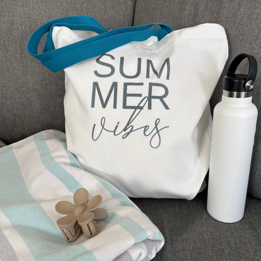 Lifestyle photo of tote bag with 'Summer Vibes' text, white water bottle, and floral hair clip on a gray surface.